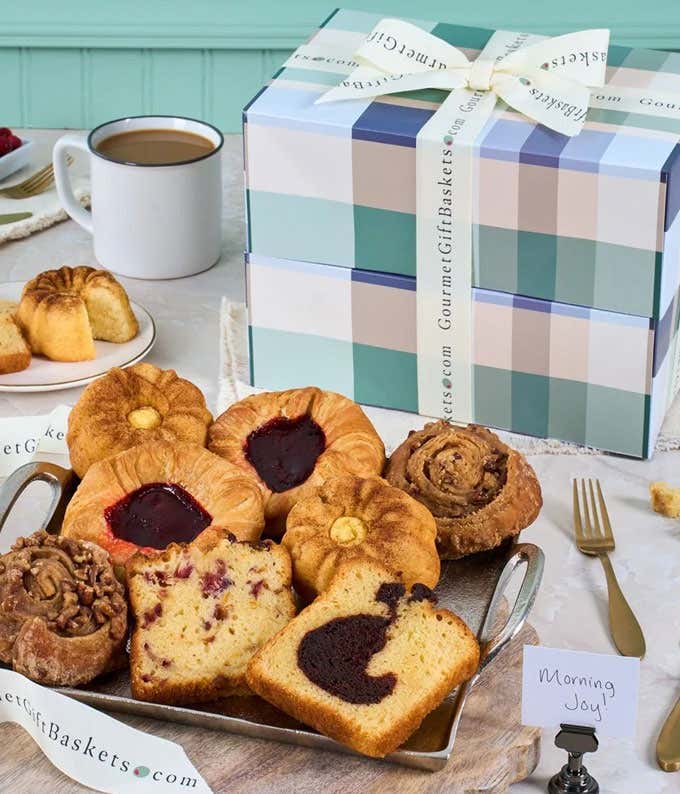 Gift tower with pastel plaid boxes filled with baked goods including cinnamon rolls, chocolate-drizzled mini cakes, marble loaf slices, cookies, and pastries, arranged on a marble surface with white plates and gold forks.
