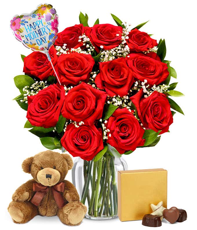 dozen red roses with greenery and baby's breath in a clear glass vase. Sat next to it is a brown teddy bear and a brown & gold square box of chocolates. And a pink and red heart shaped balloon reading happy mother's day