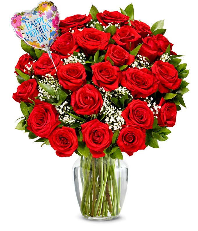 Two dozen red roses with pops of greenery and white baby's breath in a clear glass vase with a pink and red heart shaped balloon reading happy mother's day. 