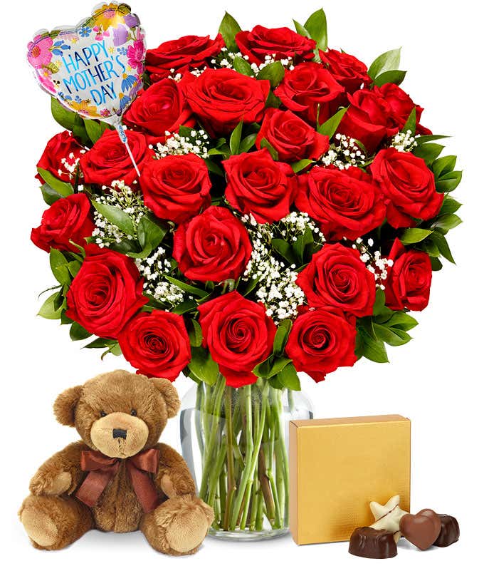 Two dozen red roses with greenery and baby's breath in a clear glass vase. Sat next to it is a brown teddy bear and a brown & gold square box of chocolates. And a pink and red heart shaped balloon reading happy mother's day