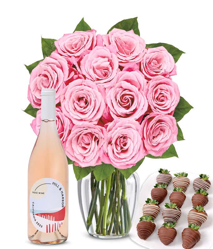 A bouquet of 1 dozen soft pink roses arranged in a clear glass vase is displayed beside a bottle of Hill & Harbor Rosé and a neat assortment of 12 chocolate-covered strawberries.