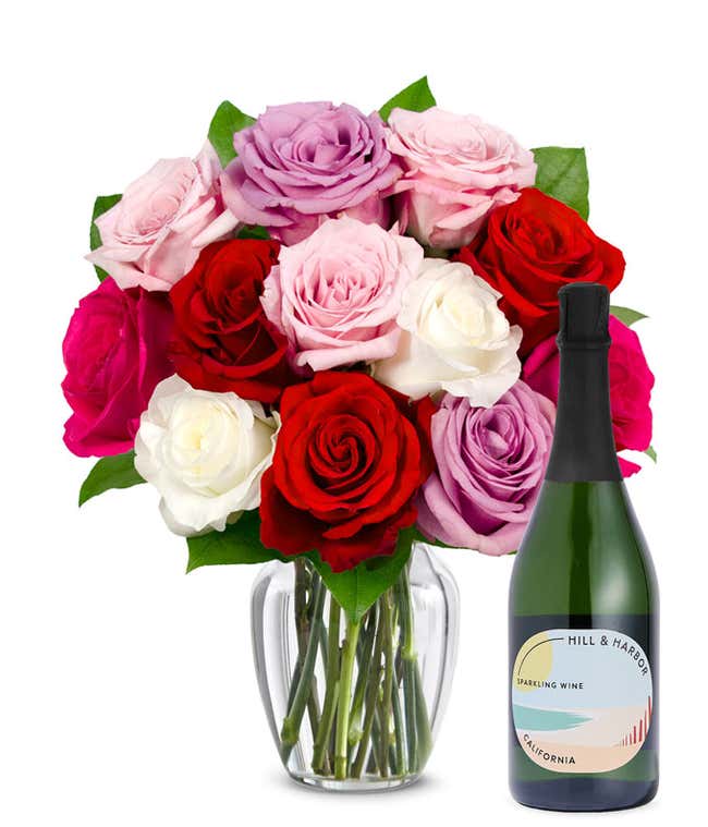 One dozen pink, purple and red roses arranged in a clear glass vase sits beside a bottle of Hill &amp; Harbor California Sparkling Wine.