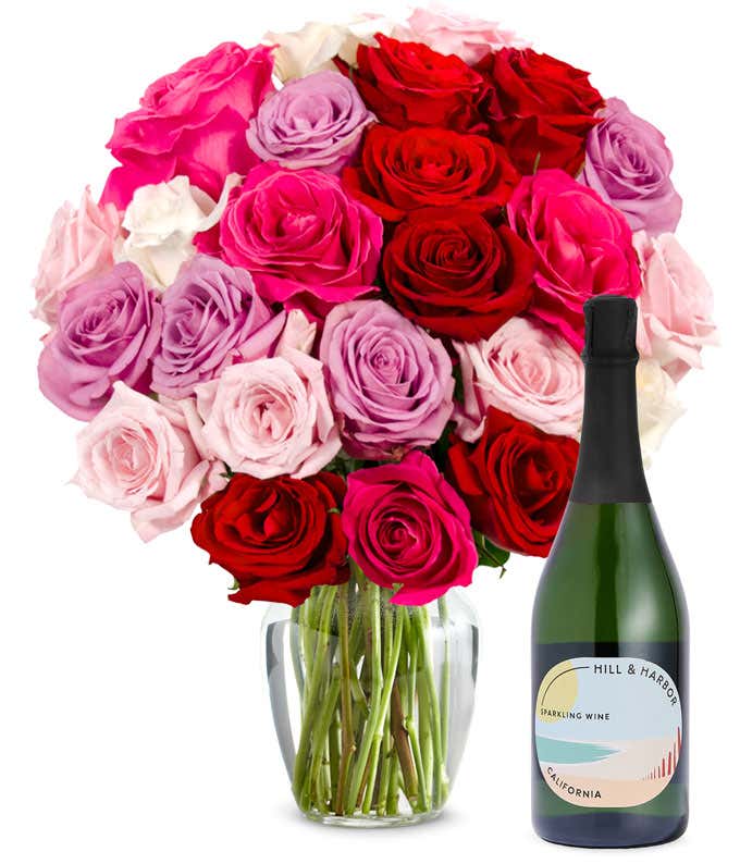Two dozen pink, purple and red roses arranged in a clear glass vase sits beside a bottle of Hill & Harbor California Sparkling Wine.