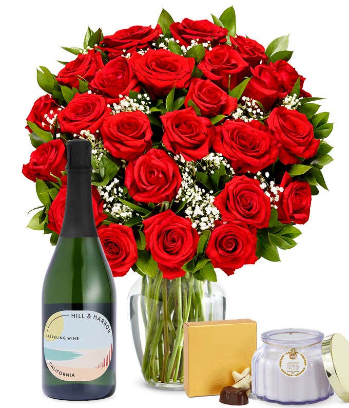 Two Dozen Red Roses in a clear glass vase with a box of chocolates, a bottle of sparkling wine, and a Dahlia & Orchid scented candle