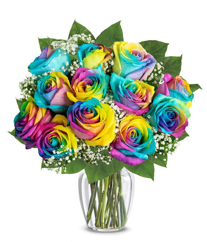 A colorful bouquet of rainbow roses in a clear glass vase, with petals in vibrant hues of blue, yellow, pink, and purple, accented by baby's breath and green leaves, creating a bright and cheerful display.