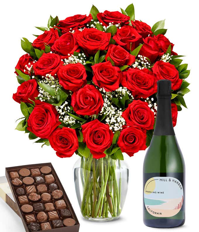 A lush arrangement of 2 dozen red roses in a clear vase sits beside a bottle of Hill & Harbor Sparkling Wine and a 1-pound box of gourmet assorted chocolates.
