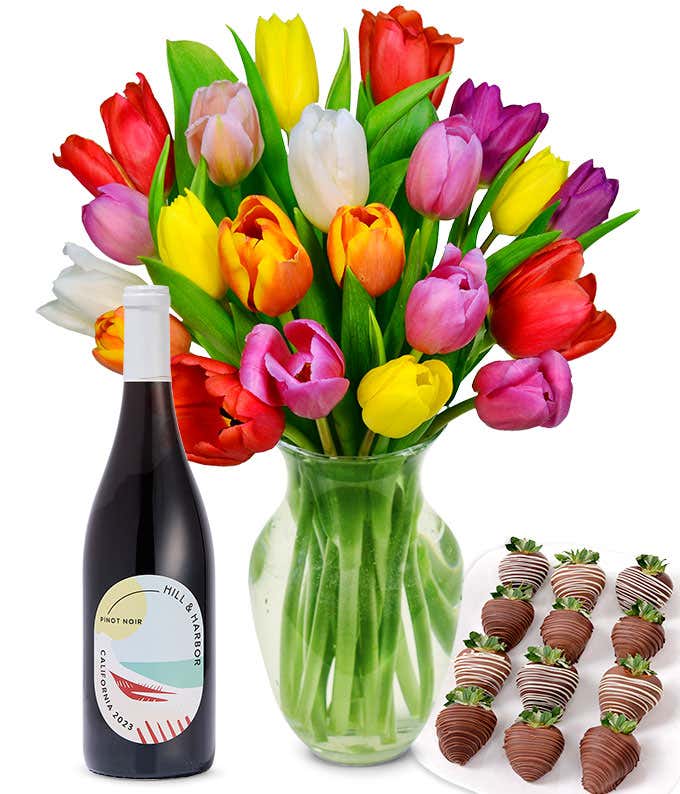 A clear glass vase filled with a 20-stem assortment of multi-colored tulips paired with a bottle of red wine and 12 chocolate-covered strawberries.