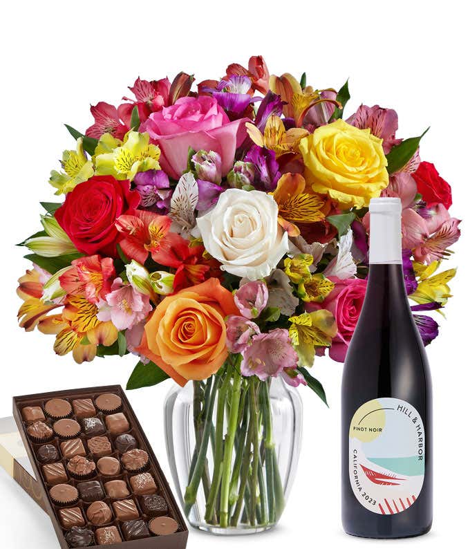 A colorful mixed bouquet of roses and alstroemeria flowers arranged in a clear vase is displayed beside a bottle of Hill & Harbor Pinot Noir and a box of assorted chocolates.