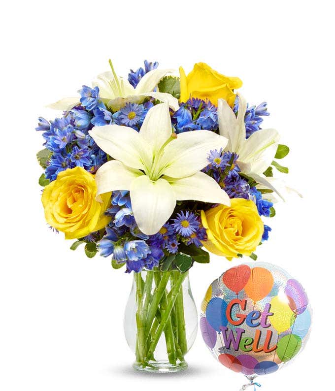 A floral arrangement featuring white Asiatic lilies, yellow roses, blue delphinium, and purple Monte Casino in a keepsake glass vase, accompanied by a 'Get Well' mylar balloon.