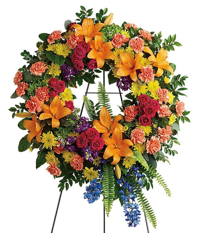 A standing funeral wreath adorned with green hydrangeas, hot pink spray roses, orange asiatic lilies, orange carnations, blue delphinium, purple stock, yellow cushion chrysanthemums, and floral greenery, symbolizing eternal grace and heartfelt remembrance