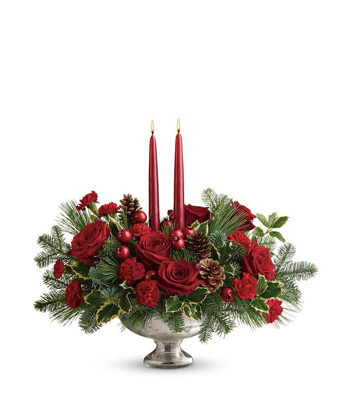 Holiday centerpiece featuring red roses, red miniature carnations, festive berry sprays, pinecones, evergreen greenery, and two red tapered candles arranged in a keepsake mercury glass bowl