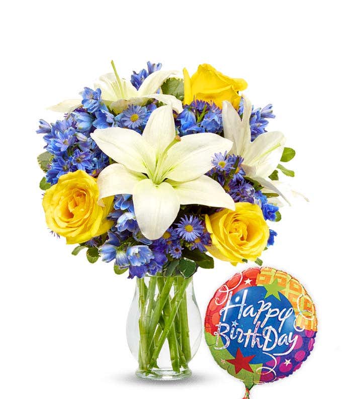 Birthday flower bouquet with white lilies, yellow roses, blue delphinium, and purple asters in a clear glass vase, paired with a colorful Happy Birthday balloon.