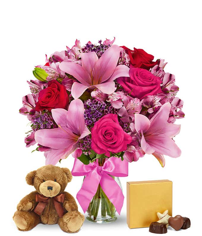 Romantic gift set with a bouquet of pink lilies, hot pink and red roses, pink alstroemeria, and purple filler in a glass vase with a pink bow, paired with a plush teddy bear and box of assorted chocolates. 