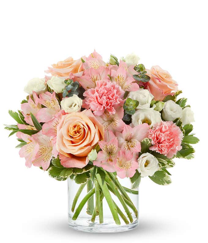 Bouquet of peach roses, pink carnations, pink alstroemeria, and white lisianthus with greenery, arranged in a clear glass cylinder vase.