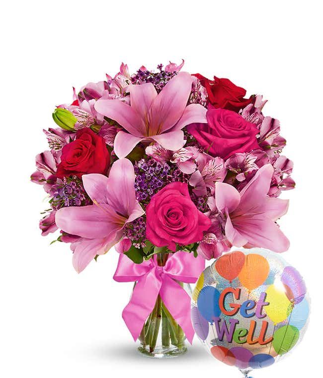 Clear glass vase filled with pink lilies, hot pink roses, red roses, pink alstroemeria, and purple filler flowers, tied with a pink bow and paired with a colorful 'Get Well' balloon. 