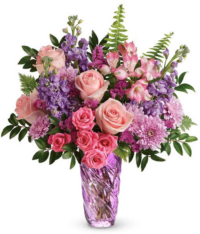 Full bouquet of pink roses, hot pink spray roses, lavender stock, pink alstroemeria, purple poms, and greenery arranged in a lavender swirl glass vase.