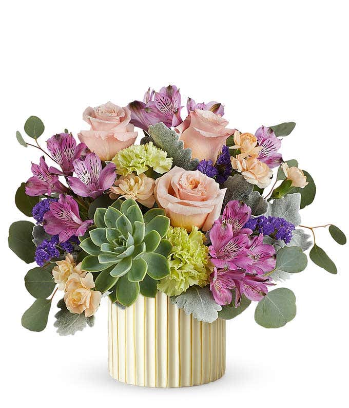 Floral arrangement with blush roses, purple alstroemeria, green carnations, small peach blooms, a large green succulent, and eucalyptus in a ribbed ivory planter with gold accents.