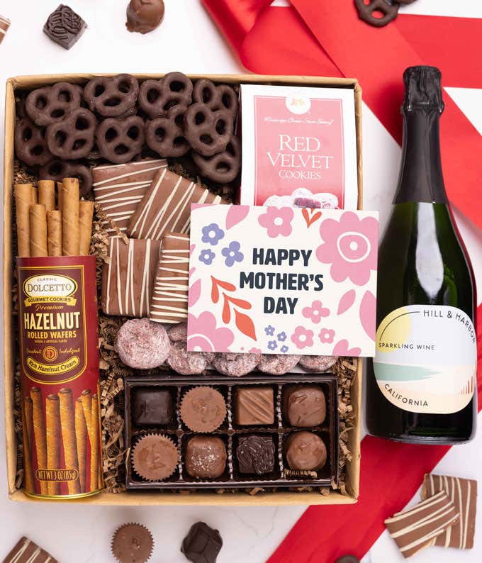 A bottle of Hill & Harbor sparkling wine, a box of assorted chocolates, chocolate-covered pretzels, and mini chocolate chip cookies with a 'Happy Mother's Day' card placed above.