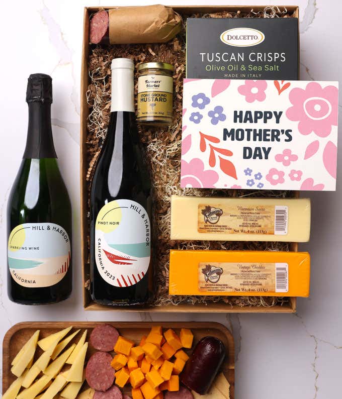 A snack and wine gift box featuring 2 bottles of wine - red and sparkling, 2 cheeses - chaddar & Swiss, cracker crisps, summer sausage, and mustard. A 'Happy Mother's Day,' sits on top. A perfect gift for Mom.