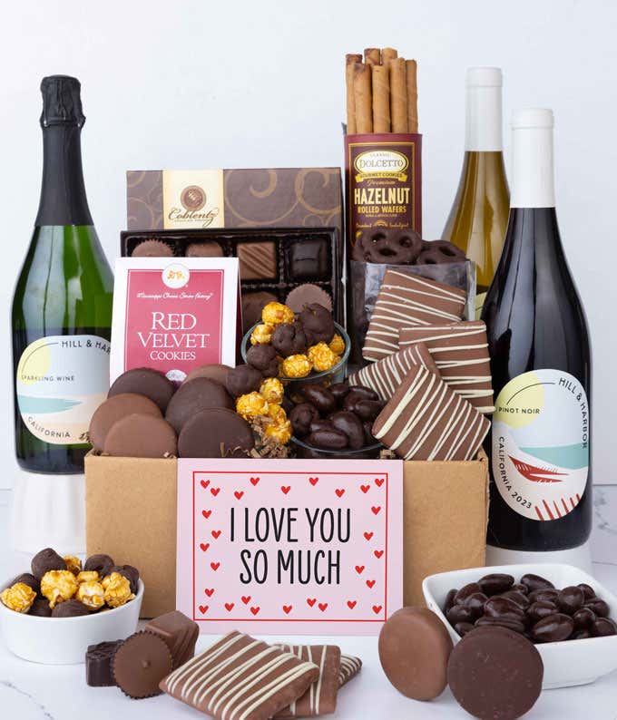 A gift box overflowing with treats including an assortment of chocolate-covered items like oreoes, almonds graham crackers and pretzels along with red velvet cookies, hazelnut wafers, an 8 piece box of chocolates, 3 bottles of wine and an I Love You card.